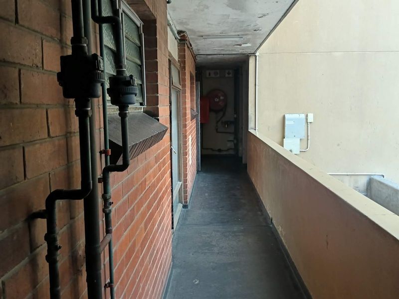 An exterior apartment access walkway featuring exposed brickwork, visible plumbing infrastructure, and a fire hose reel, showing signs of significant wear and aging.