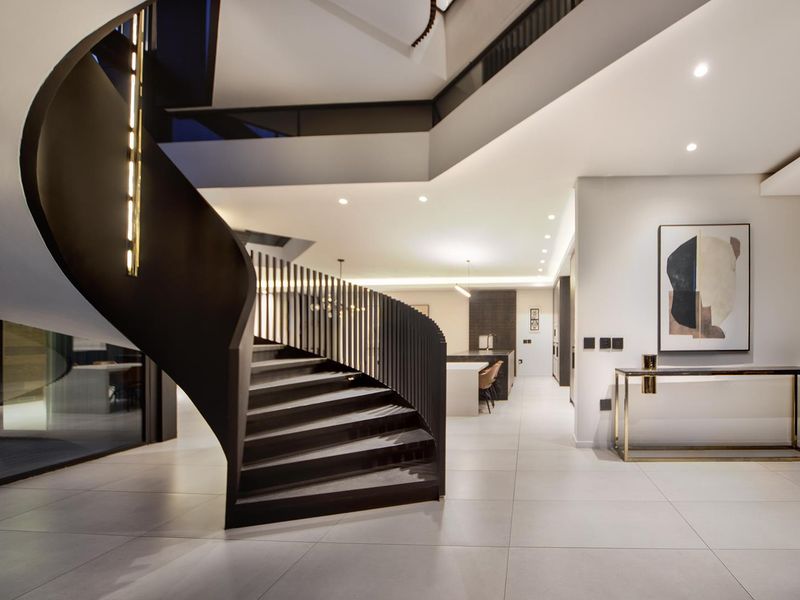 Modern entrance hall featuring a striking curved staircase, high ceilings, and sleek tiled flooring, leading into an open-plan living and kitchen area.