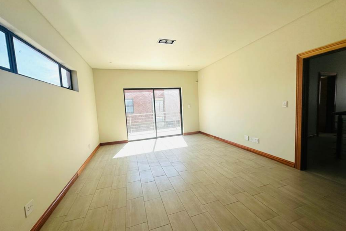 A bright, empty living room featuring tiled flooring, recessed ceiling lighting, and a sliding glass door leading to an outdoor balcony.