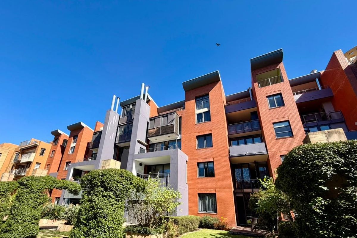A low-angle exterior view of a modern multi-story brick apartment complex featuring balconies and landscaped garden areas.