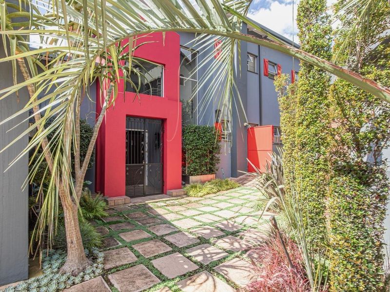 Front entrance area featuring a bold red architectural facade, secure metal gate, and a well-maintained paved walkway surrounded by landscaped garden beds.