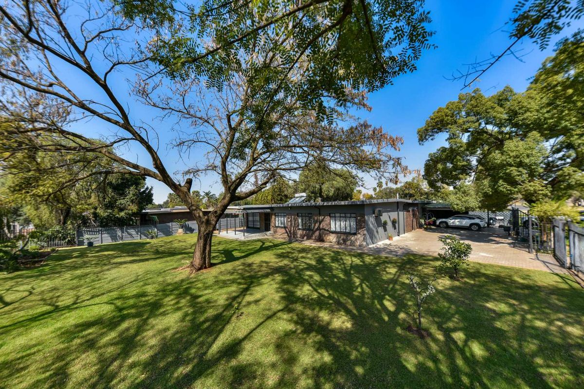 A spacious rear garden featuring a well-maintained lawn, mature trees, a paved driveway, and a covered patio area attached to the main house.