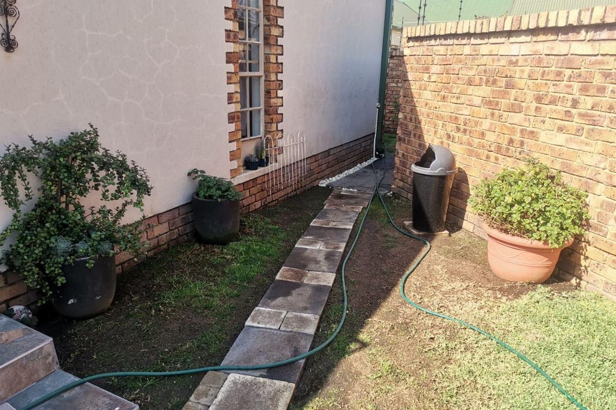 A side garden area featuring a paved walkway, brick boundary wall, and plastered exterior wall with brick detailing.