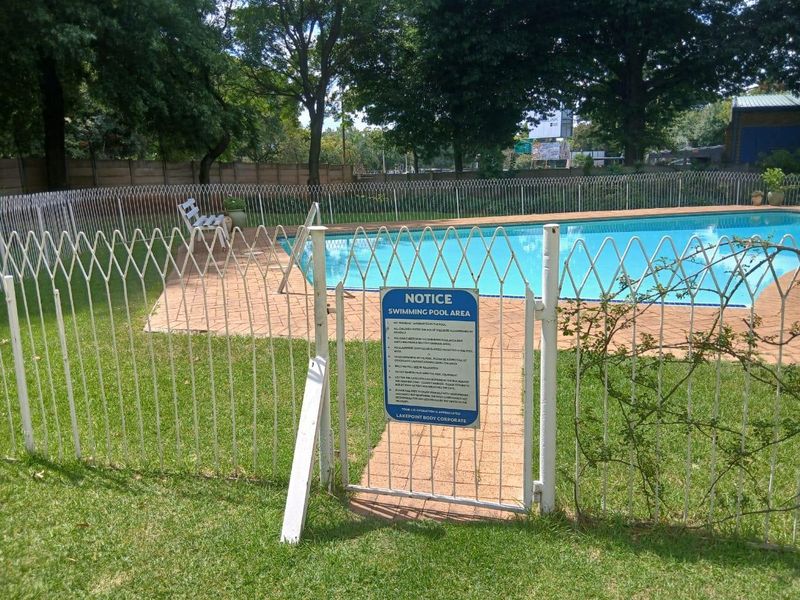 A communal swimming pool area featuring paved surrounds, manicured lawn, and secure metal perimeter fencing with a gated entrance.