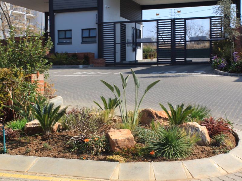 A well-maintained residential complex entrance featuring a secure gated access point, guard house, and landscaped garden beds.