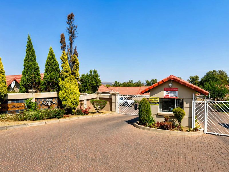 The entrance to the townhouse complex features a secure guard house, gated access, and a paved driveway with established landscaping.