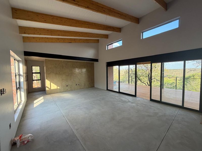 Spacious open-plan living area featuring polished concrete floors, exposed timber beams, and large sliding doors opening onto a wooden deck with scenic views.