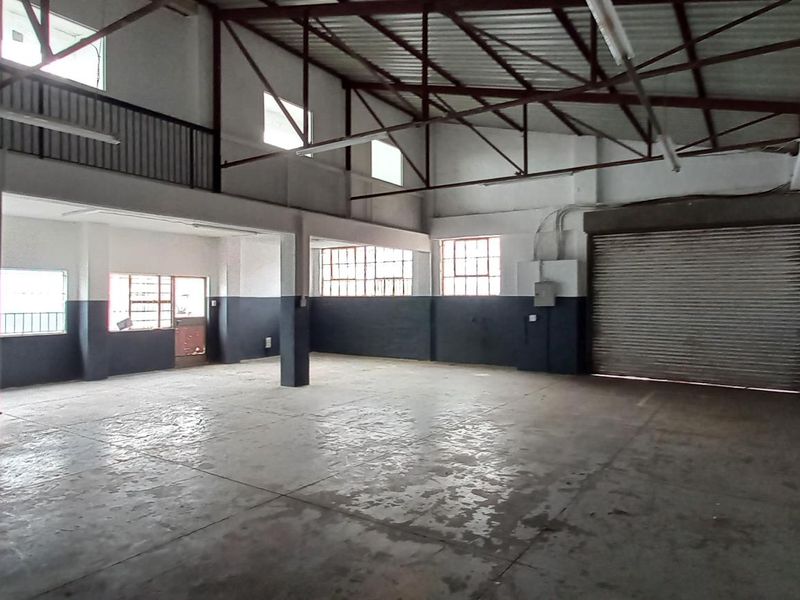 Spacious industrial utility area featuring high ceilings with exposed steel trusses, a large roller shutter door, concrete flooring, and a mezzanine level.