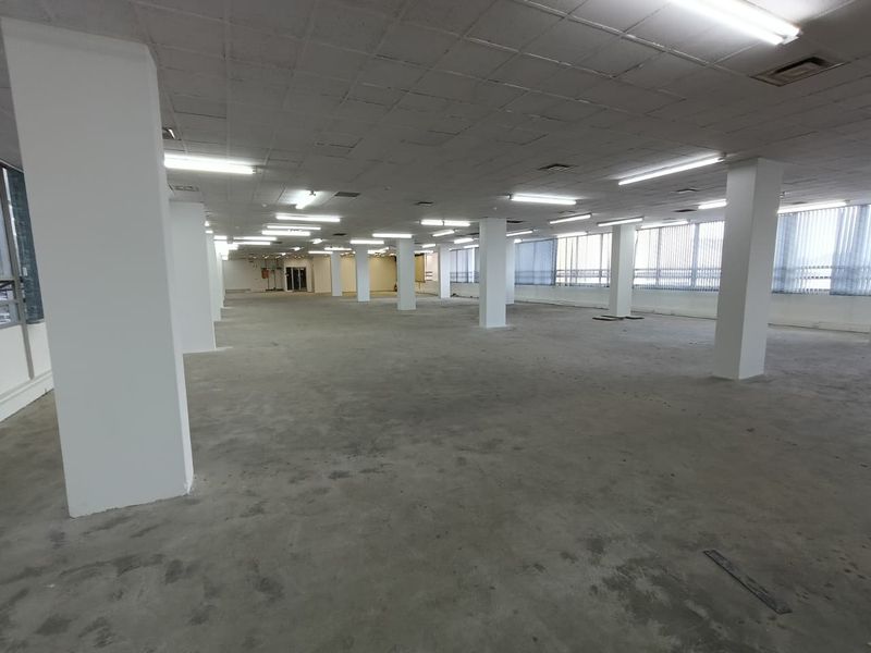 A large, open-plan office space featuring structural columns, suspended ceilings with fluorescent lighting, and expansive windows with vertical blinds.