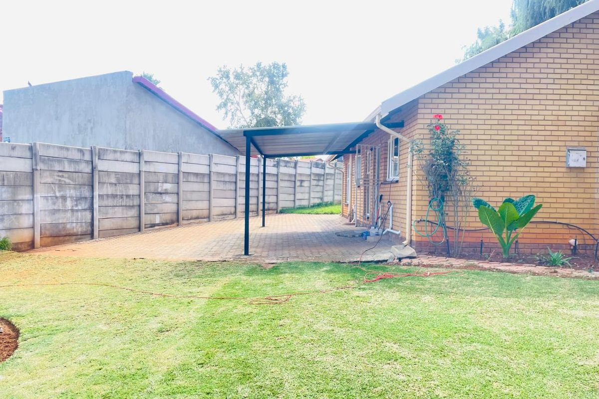 A rear view of the property featuring a paved patio area with a metal-framed cover, adjacent to a brick exterior and a lawned garden.