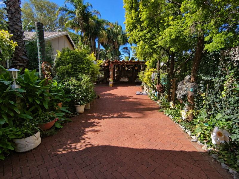 A lush, well-established garden featuring a brick-paved pathway leading toward a covered outdoor patio area surrounded by mature trees and greenery.
