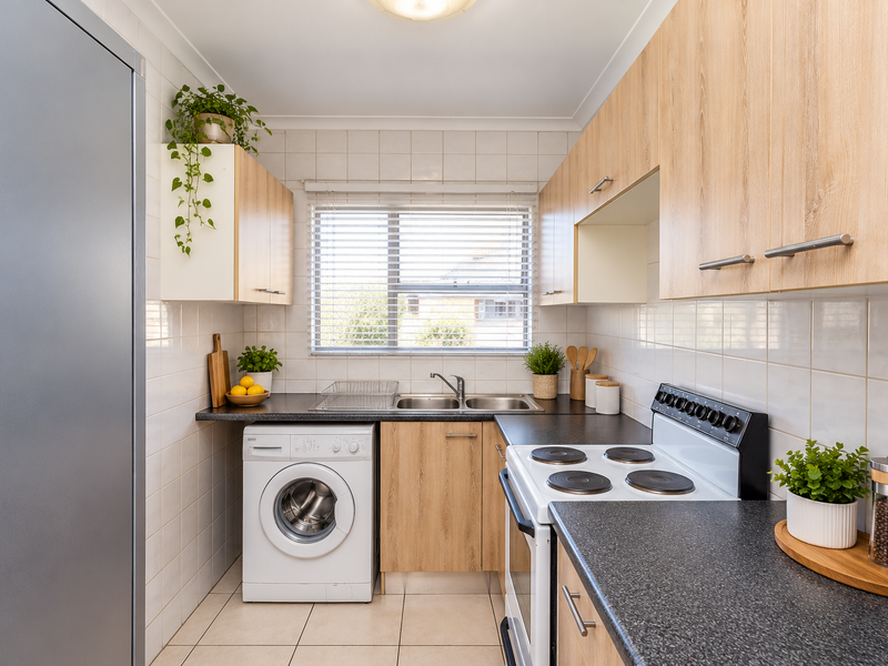 A functional kitchen featuring light wood-tone cabinetry, dark laminate countertops, a stainless steel sink, and a built-in electric stove and washing machine.