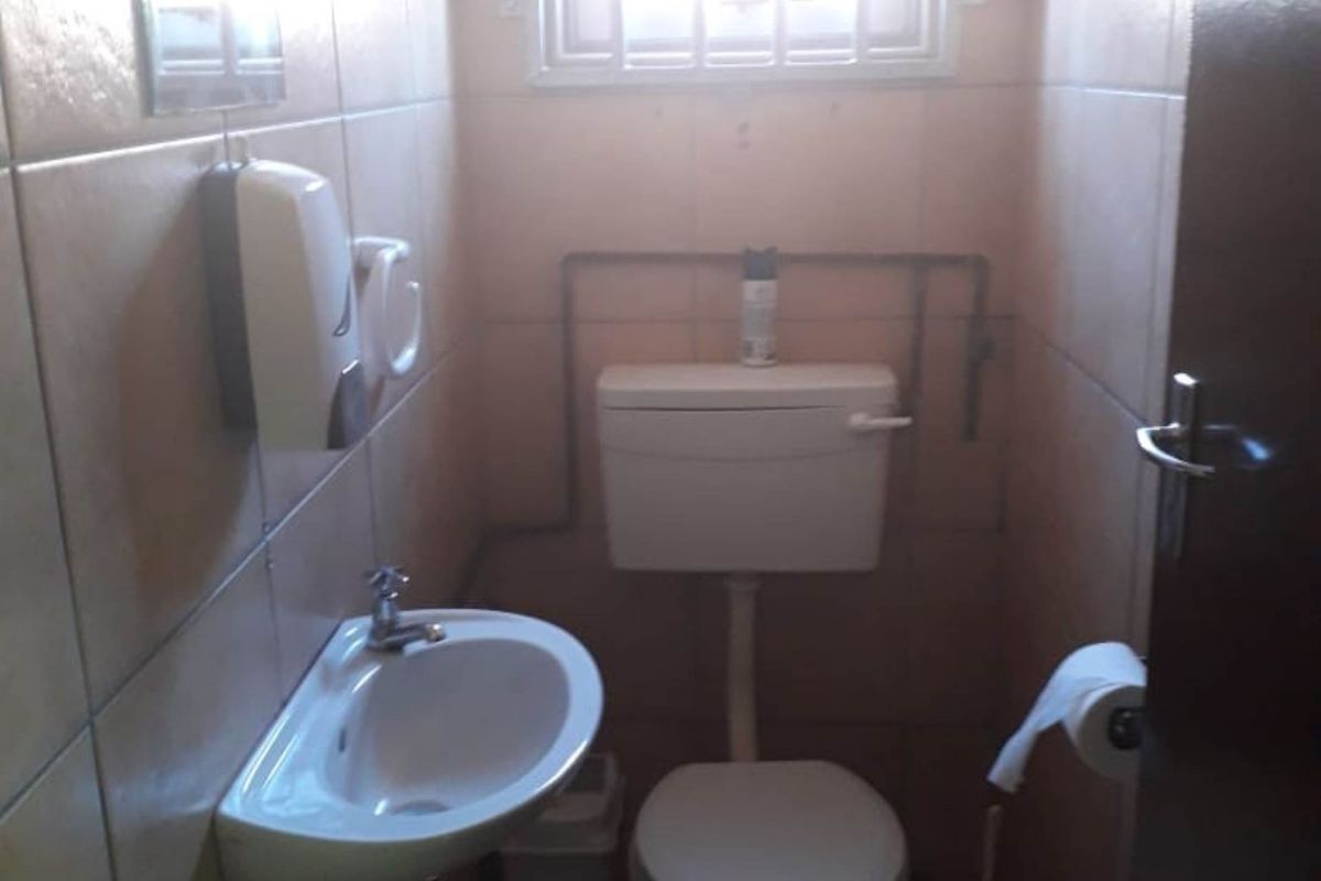 A compact industrial-style restroom featuring a wall-mounted basin, standard toilet, and a window with security bars.