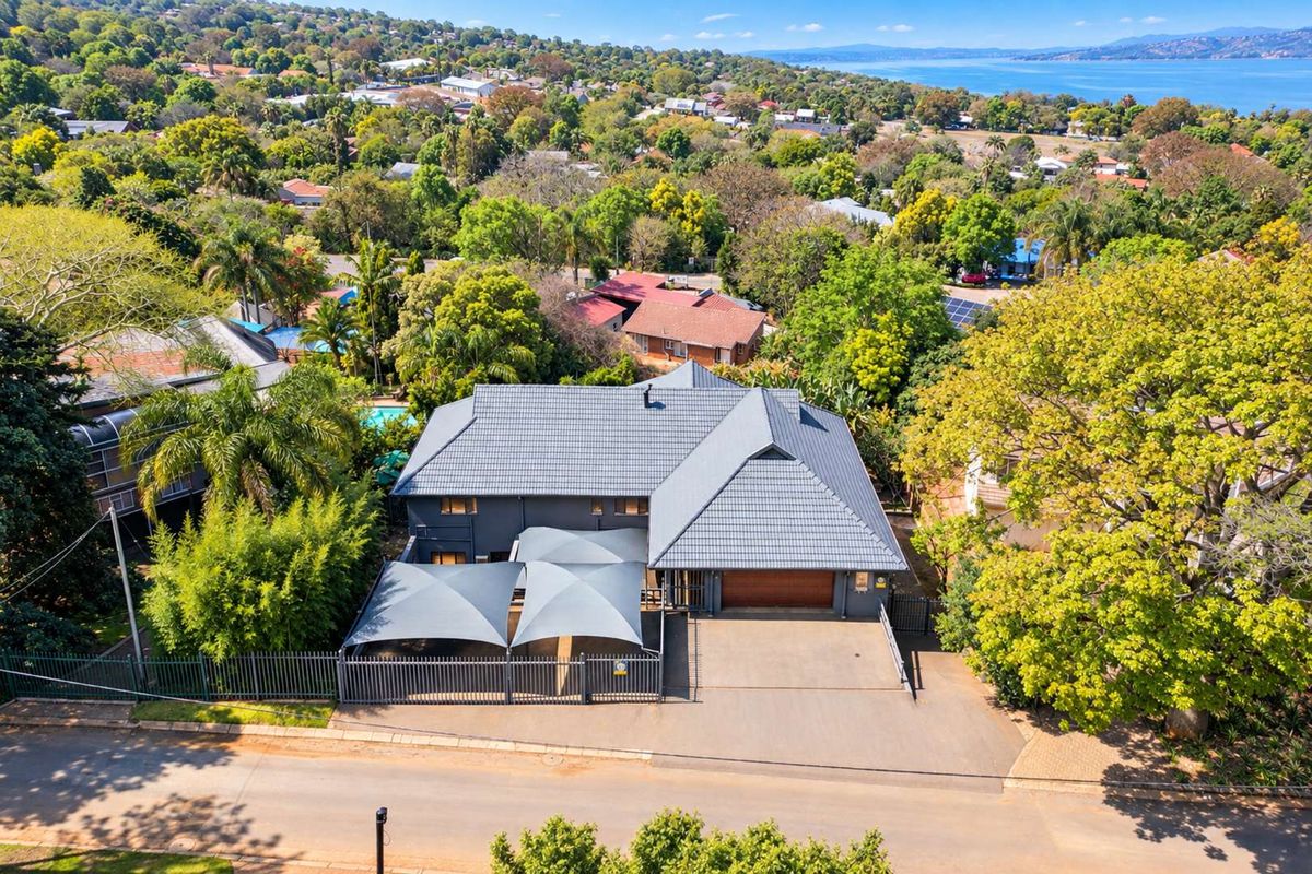 Aerial view of a residential property featuring a tiled roof, paved driveway, garage, and a shaded parking area within a landscaped suburban setting.