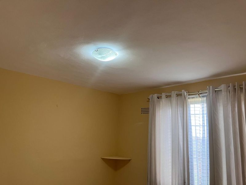 A simple, empty bedroom featuring yellow walls, a ceiling light fixture, and a built-in corner shelf near the window.