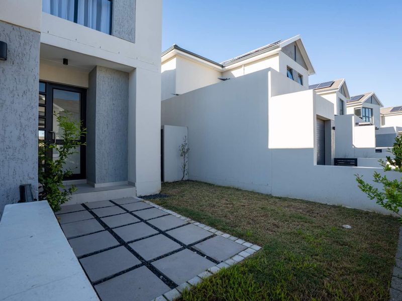A well-maintained rear exterior view of the townhouse featuring a paved patio area, a small lawn, and modern boundary walls.