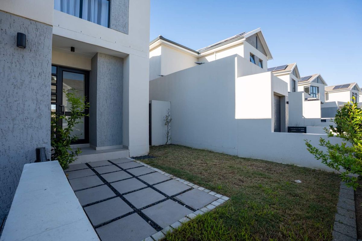 A well-maintained rear exterior view of the townhouse featuring a paved patio area, a small lawn, and modern boundary walls.