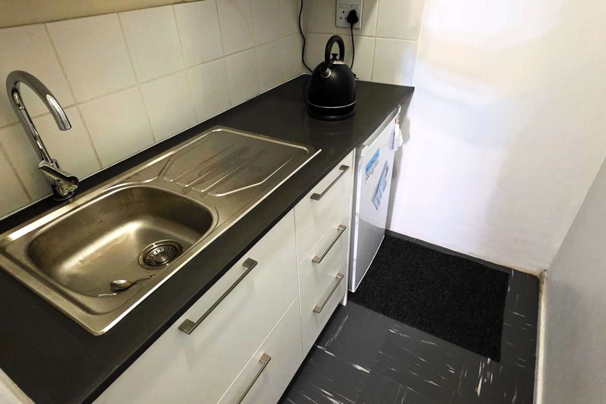 A compact kitchenette featuring a stainless steel sink, dark laminate countertop, white cabinetry, and tiled splashback, suitable for an office break area.