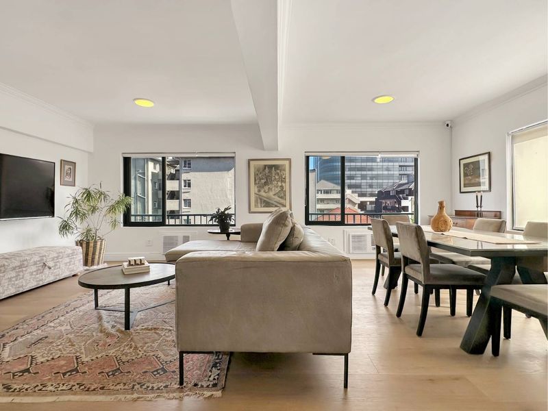Spacious open-plan living and dining area featuring light timber flooring, recessed lighting, and large black-framed windows with urban views.