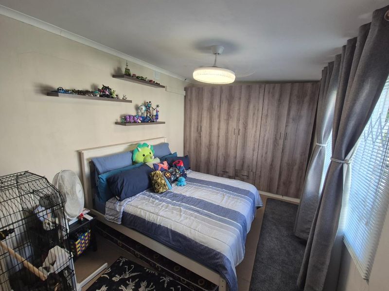 A well-maintained bedroom featuring extensive built-in wooden cupboards, neutral wall finishes, and large windows with fitted blinds and curtains.