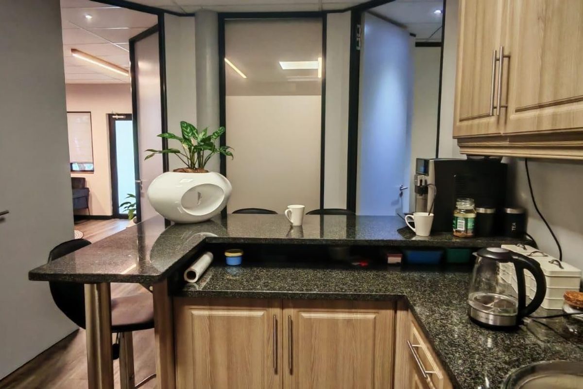A compact office kitchenette featuring granite countertops, light wood-finish cabinetry, and a breakfast bar, integrated into a modern workspace with glass partition doors.