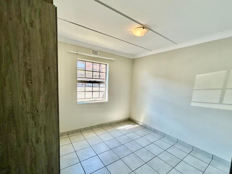 A bright, empty bedroom featuring tiled flooring, a built-in cupboard, and a window with security bars, offering a clean and neutral space.