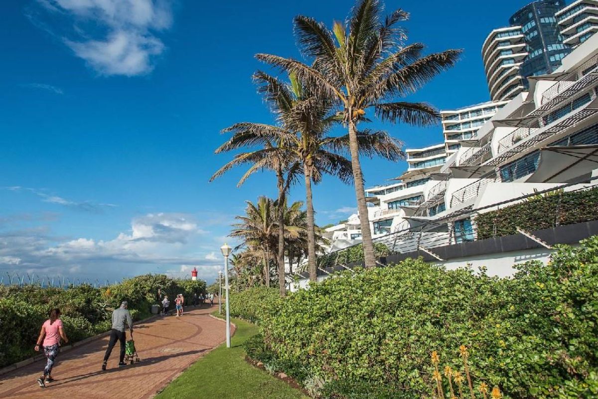 A scenic view of the apartment complex exterior, featuring landscaped gardens, paved walkways, and proximity to the coastal promenade.