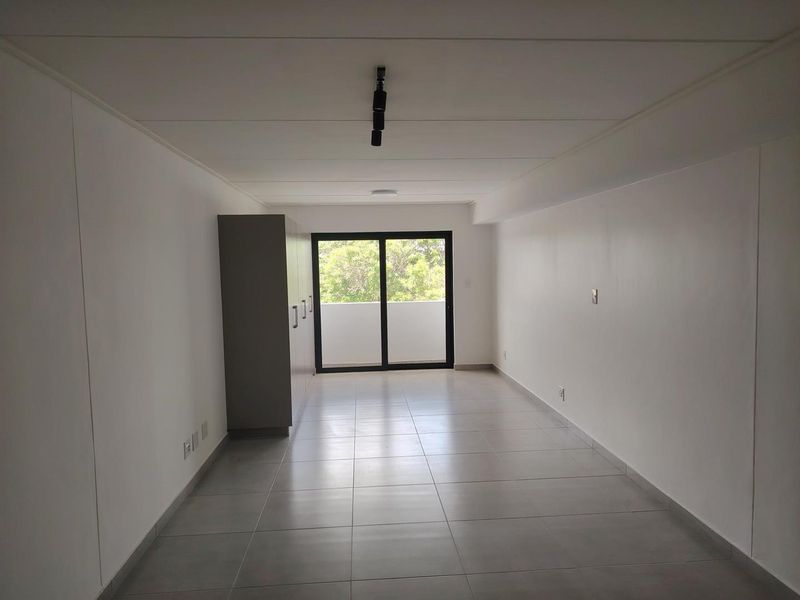 A bright, empty bedroom featuring neutral tiled flooring, built-in cupboards, and a sliding glass door leading to an outdoor balcony area.