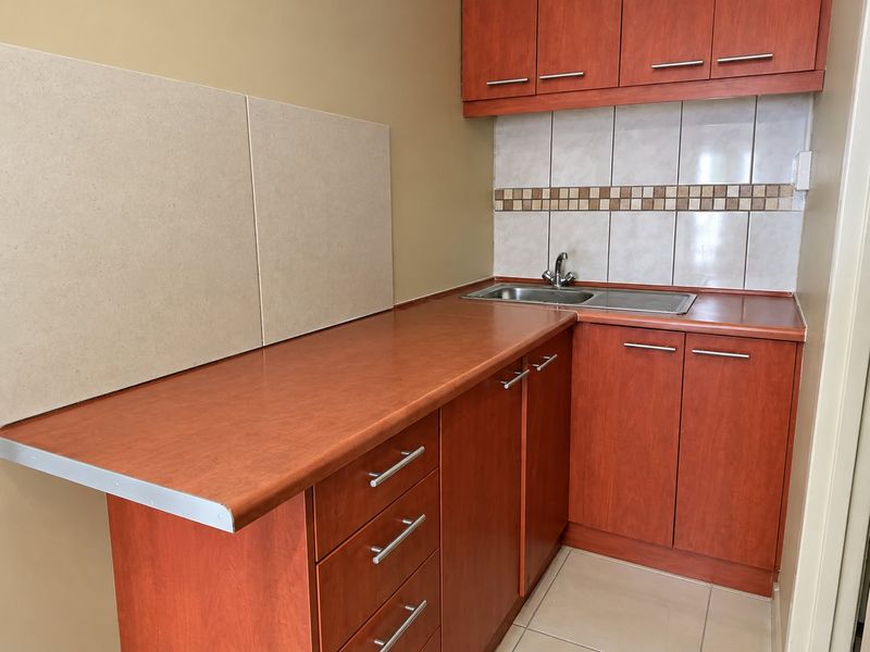A compact kitchen featuring wood-look laminate cabinetry, tiled backsplash, stainless steel sink, and neutral floor tiles, offering a functional and clean preparation space.