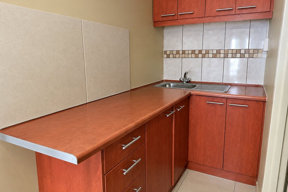 A compact kitchen featuring wood-look laminate cabinetry, tiled backsplash, stainless steel sink, and neutral floor tiles, offering a functional and clean preparation space.