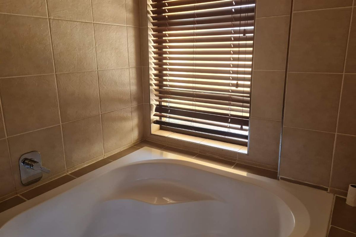 A bathroom corner featuring a built-in soaking tub with tiled surround and a window fitted with wooden venetian blinds.