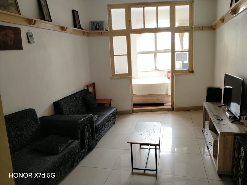 A living room featuring tiled flooring and wooden wall shelving, with a view through a multi-pane window frame into an adjacent bedroom.