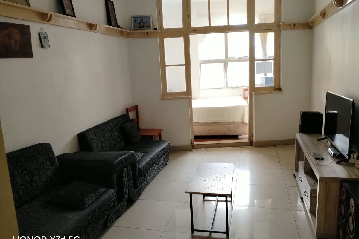 A living room featuring tiled flooring and wooden wall shelving, with a view through a multi-pane window frame into an adjacent bedroom.