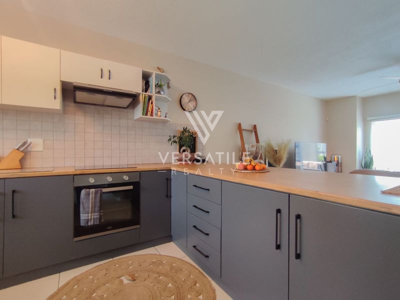 Modern open-plan kitchen featuring grey cabinetry, light wood-tone countertops, built-in oven, extractor fan, and a tiled backsplash, seamlessly flowing into the living area.