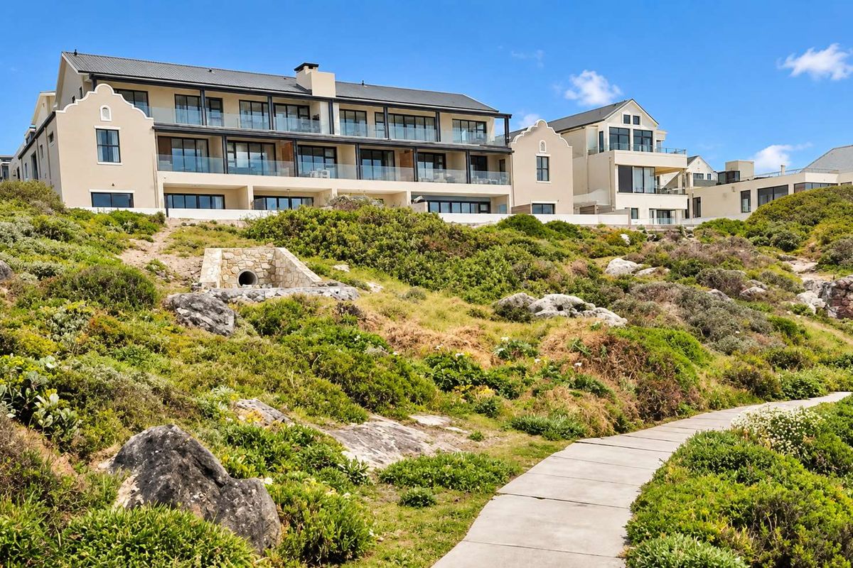 A view of the rear elevation of a multi-story apartment complex featuring balconies and a paved walkway through landscaped coastal vegetation.