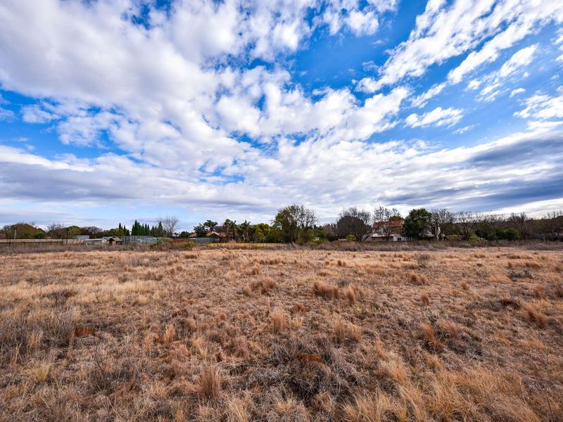 A wide-angle view of a vacant residential plot featuring dry grass and surrounding neighborhood trees and fencing.