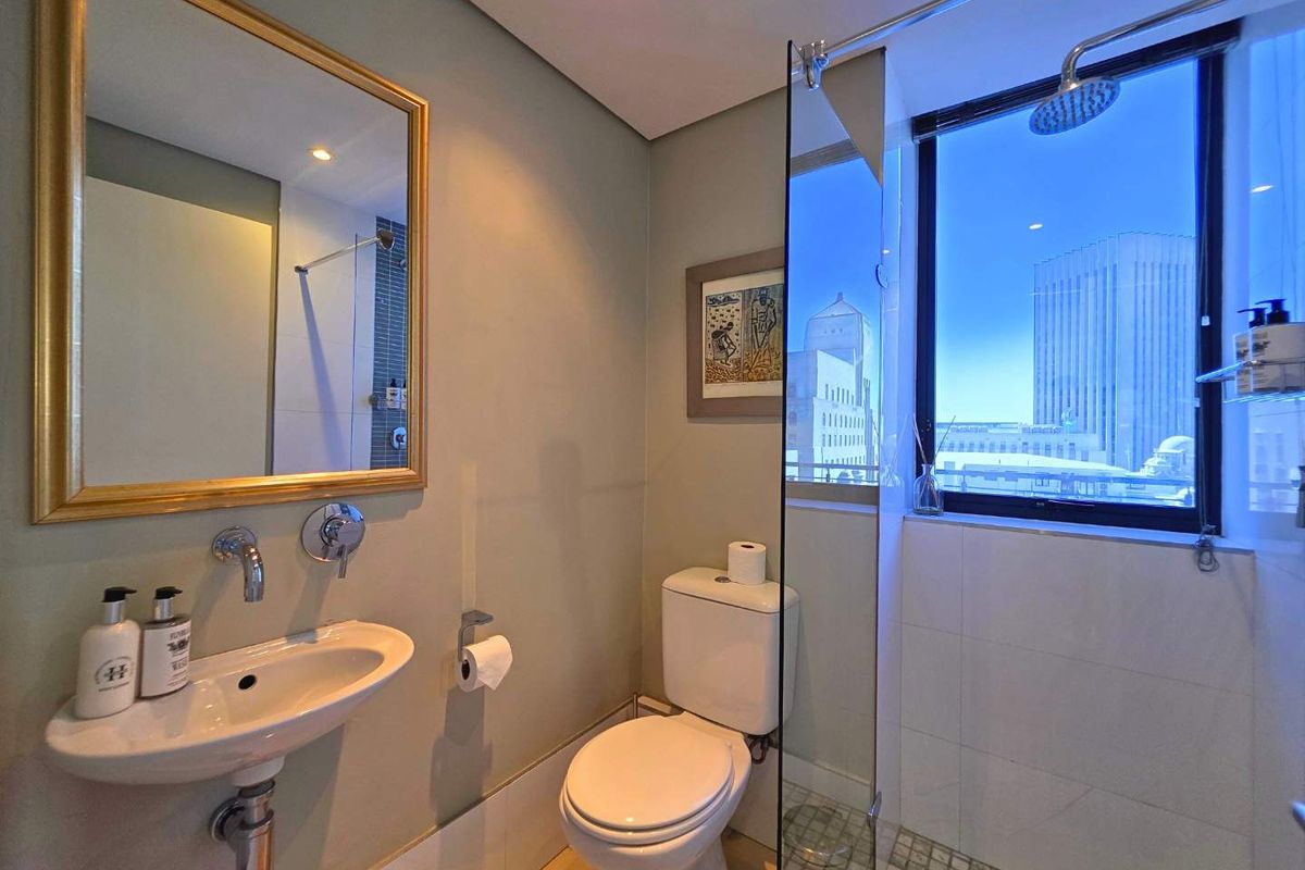 Modern bathroom featuring a glass-enclosed shower, wall-mounted basin, and toilet, complemented by a large window offering urban city views.
