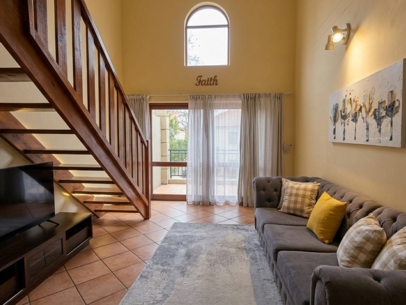 A bright living room featuring a wooden staircase, terracotta flooring, and an arched window, with direct access to a balcony through sliding glass doors.