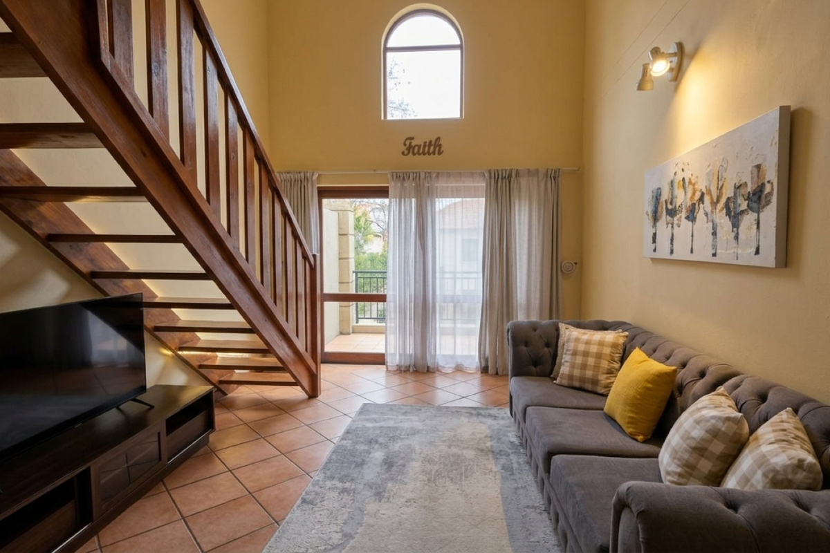 A bright living room featuring a wooden staircase, terracotta flooring, and an arched window, with direct access to a balcony through sliding glass doors.