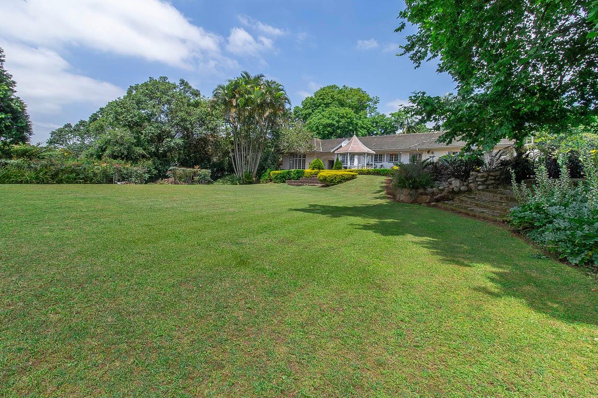 A spacious, well-maintained rear garden featuring a large manicured lawn, mature trees, and a view of the house exterior with a gazebo-style structure.