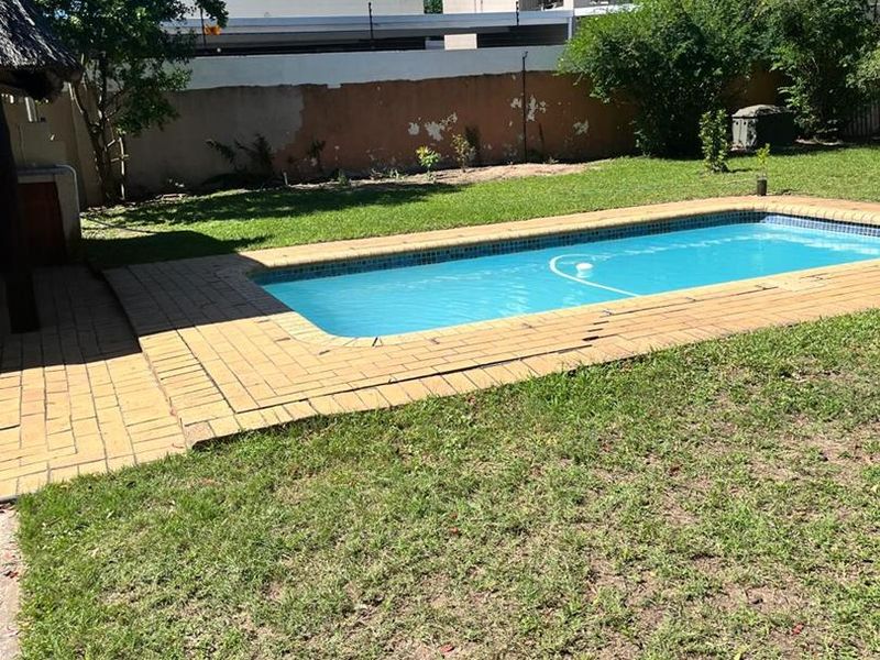Outdoor pool area featuring a swimming pool with paved surround, a thatched lapa, and a boundary wall in a private garden setting.