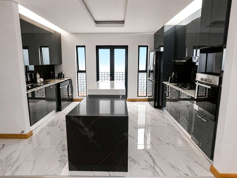 Modern kitchen featuring high-gloss black cabinetry, a central stone-topped island, integrated appliances, and elegant marble-look tiled flooring with recessed lighting.
