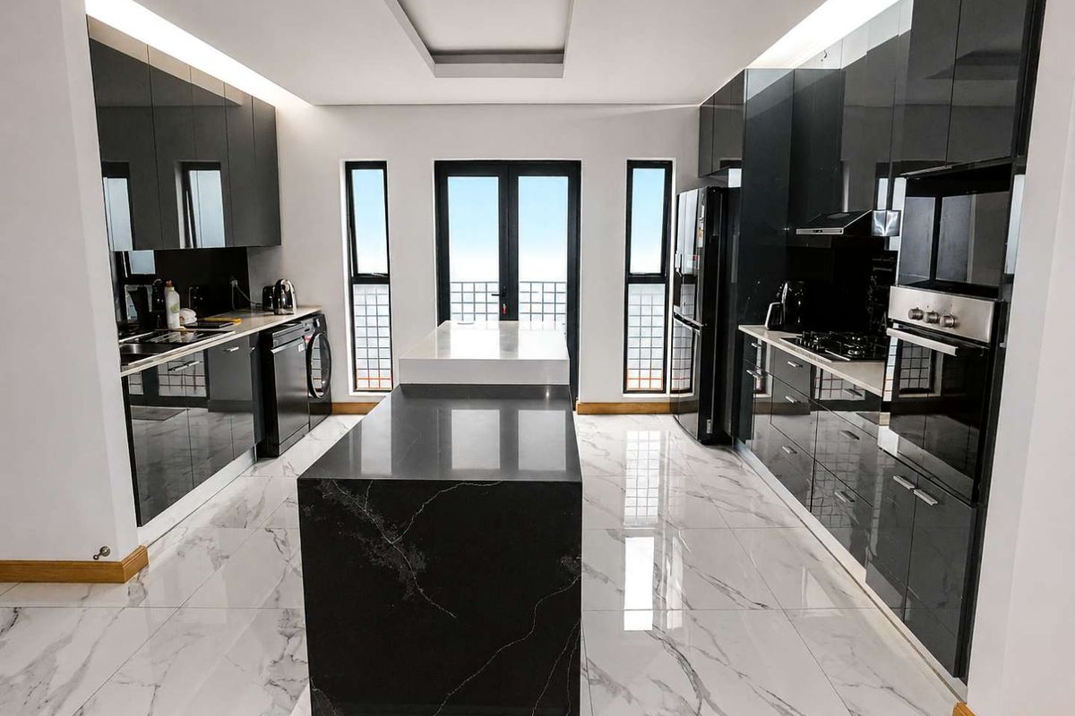 Modern kitchen featuring high-gloss black cabinetry, a central stone-topped island, integrated appliances, and elegant marble-look tiled flooring with recessed lighting.