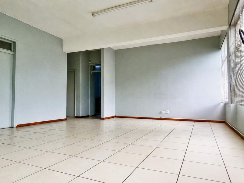 A clean, vacant office space featuring neutral tiled flooring, large windows with venetian blinds, and functional overhead lighting.