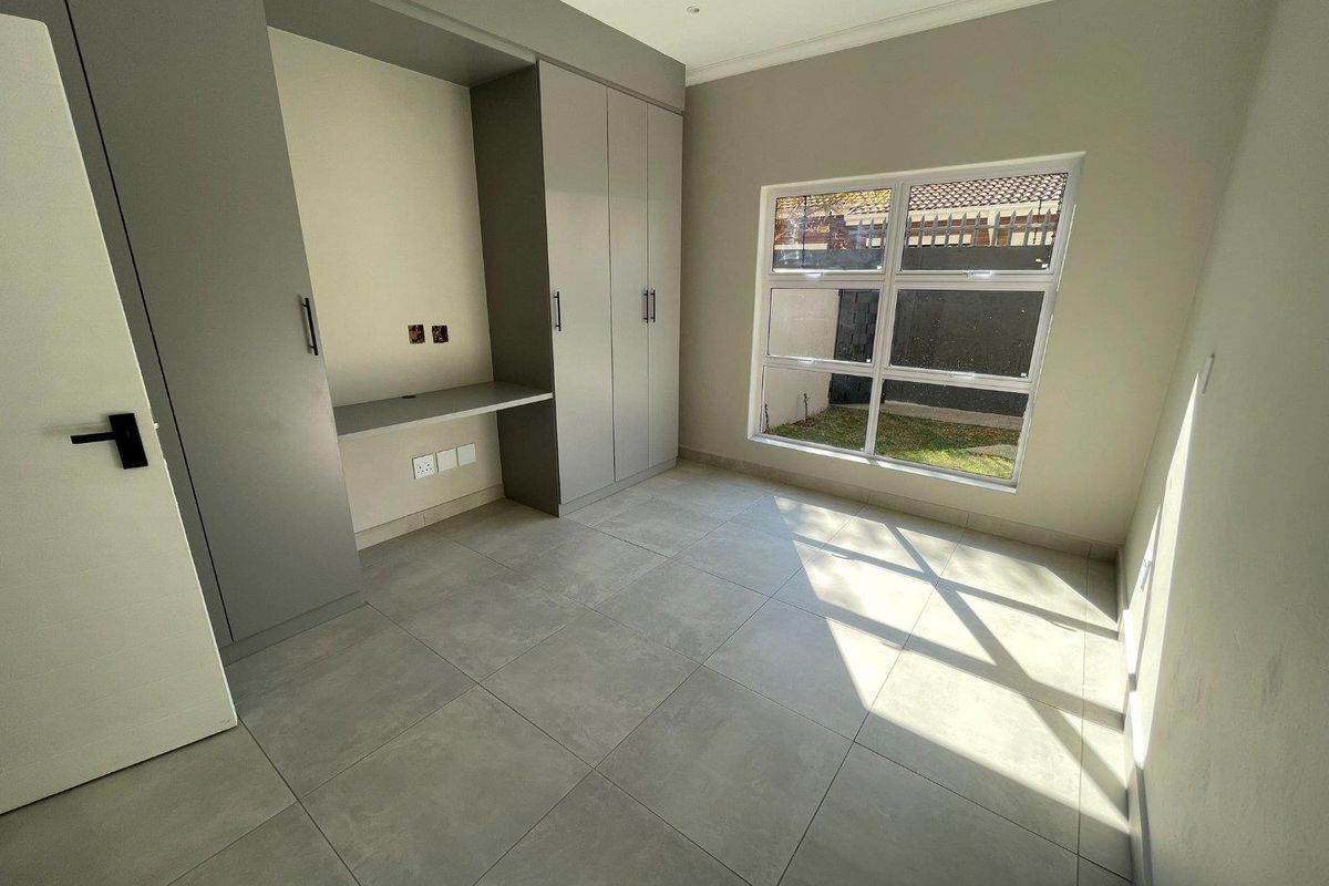 A modern, empty bedroom featuring neutral grey tiled flooring, built-in cupboards with an integrated desk, and a large window overlooking the garden.
