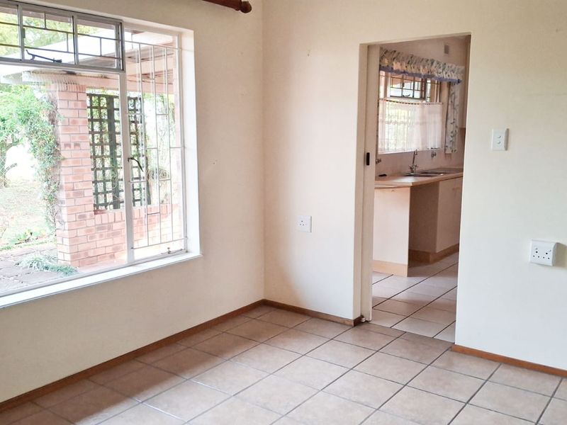 An empty living room featuring tiled flooring and a large window with security bars, providing a view of the garden and access to the kitchen.