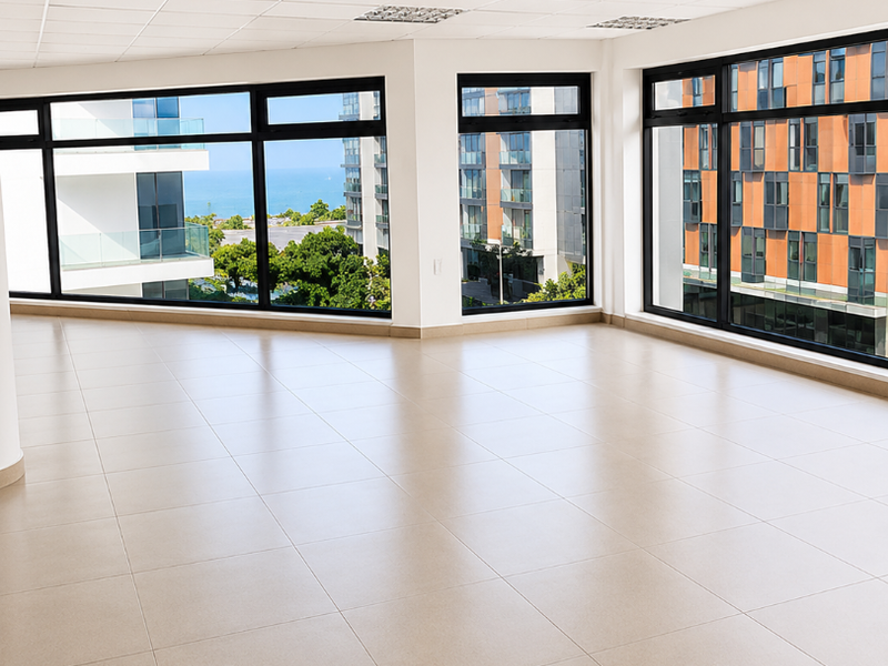 A bright, modern, empty office space featuring large windows with urban and sea views, tiled flooring, and a professional suspended ceiling.