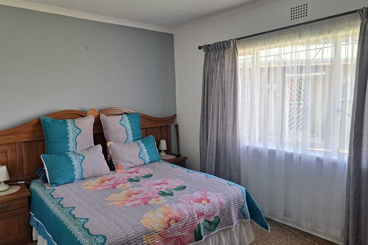 A well-lit bedroom featuring neutral wall tones, carpeted flooring, and a large window with security bars, providing a comfortable and functional living space.