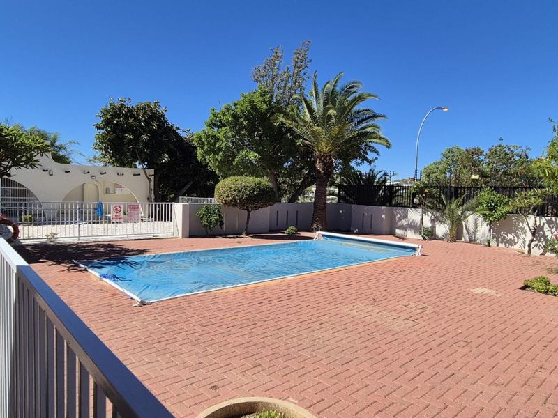 Outdoor pool area featuring a rectangular swimming pool with a cover, surrounded by extensive brick paving and secure perimeter fencing.