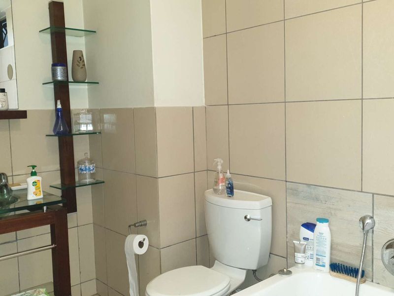 A functional bathroom featuring a glass vanity, wall-mounted shelving, a standard toilet, and a bathtub with chrome fixtures and neutral wall tiling.